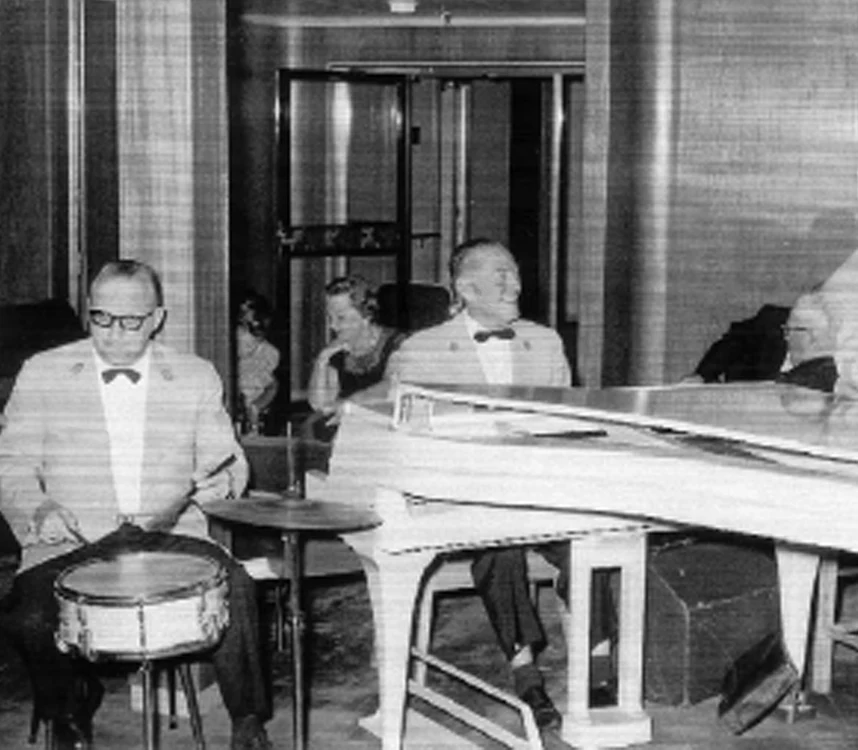Black-and-white archive photograph of a grand piano, a drummer and people indoors