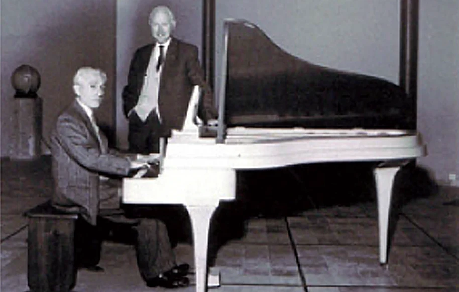 Archive photograph of one person seated at a white grand piano and another standing beside it